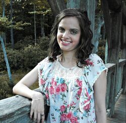 Picture of smiling White woman with shoulder-length brown hair, wearing a flowered shirt, resting her arm on the side of a wooden bridge with a forest in the background