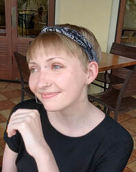 Picture of a White person with short blonde hair and a blue headband and a nose piercing, smiling and looking to the side