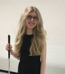Picture of woman with long blonde hair and a black dress, wearing glasses and holding a cane for blind people