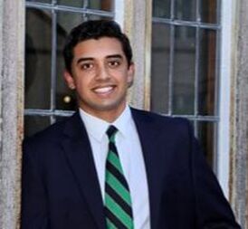Picture of man with brown skin smiling, wearing a suit and green and blue striped tie, standing in front of windows