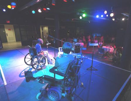Picture of man in wheelchair speaking into mic in a small theater full of people.  The stage is bathed in purple lights