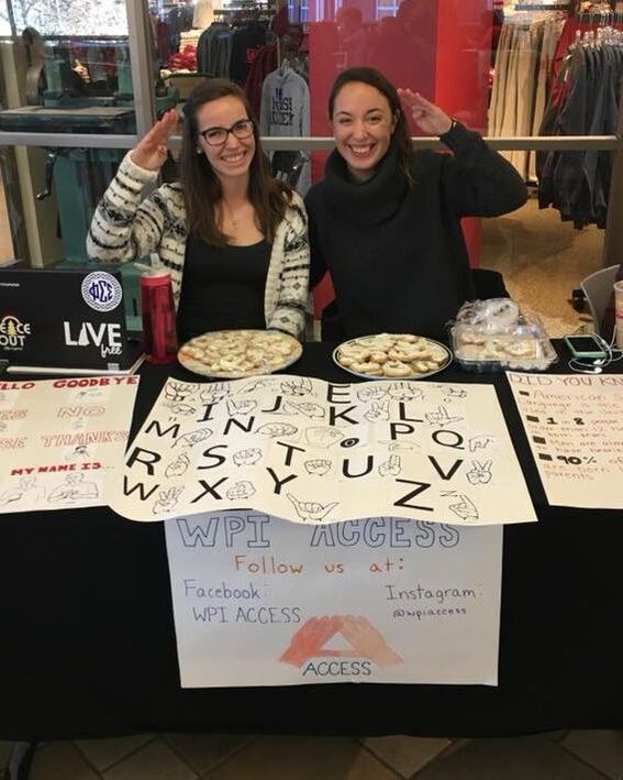 Picture of two women sitting behind a table with information about ASL and cookies, signing Hello in ASL (a wave that looks like a casual military salute).  One woman appears White, with glasses, long brown hair, and a striped sweater.  The other woman appears Asian or White, with long brownish black hair and a black hoodie.
