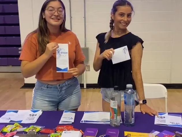 Two women holding DREAM brochures behind a table covered in DREAM information.  One woman has glasses, appears Asian, has an orange shirt and jean shorts, and long brown hair.  The other woman appears White or has light brown skin.  She has purple hair in a braid and is wearing a black shirt and jean shorts.  Both are smiling
