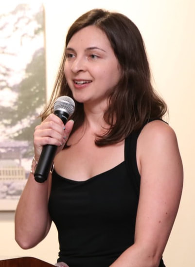 Headshot of Ashley Holben, a white woman with medium-length dark hair. She is talking into a microphone and wearing a sleeveless black dress.