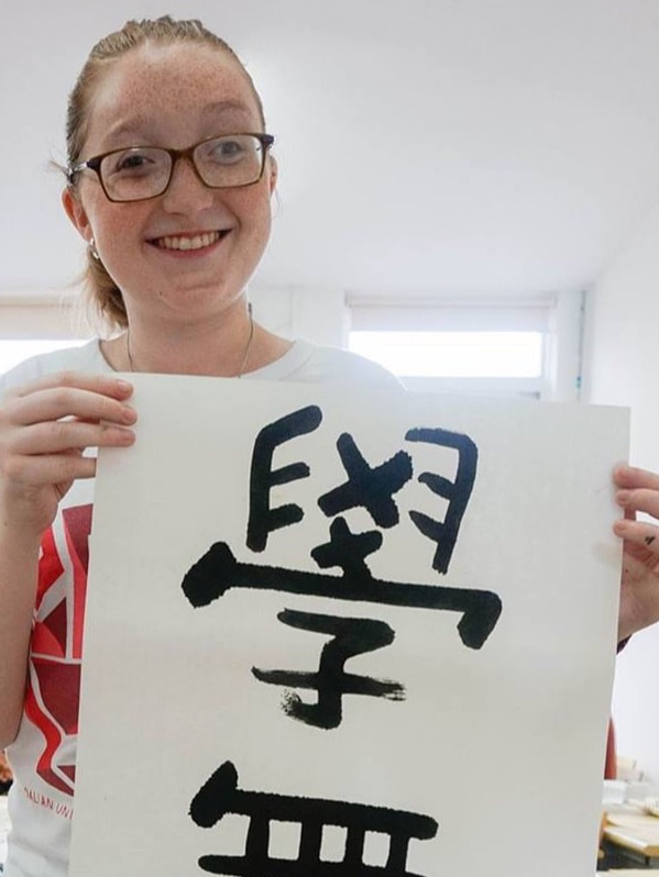 A headshot of Stephanie Collins, a white woman with her dark blonde hair tied back and wearing dark-rimmed glasses. Stephanie is holding up a large and long piece of paper with large Chinese characters painted  in black  on it.