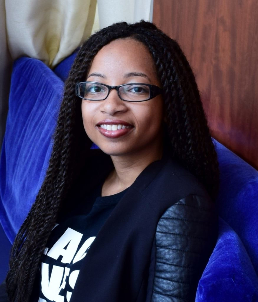 A light-skinned Black woman with long black hair and dark brown eyes, wearing dark rectangular shape glasses. Keri is smiling and wearing a Black Lives Matter tee and sitting on a blue velvet looking chair.