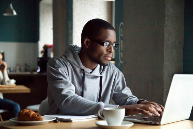 Picture of 20-something African American man sitting at a coffee shop table, typing on a laptop.  The man has glasses and a gray hoodie, and there is a croissant, a cup of coffee, and a notebook next to him on the table.