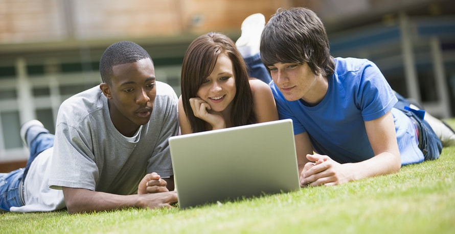 Three young college students lay on their stomachs on the grass, all looking at an open laptop. 
