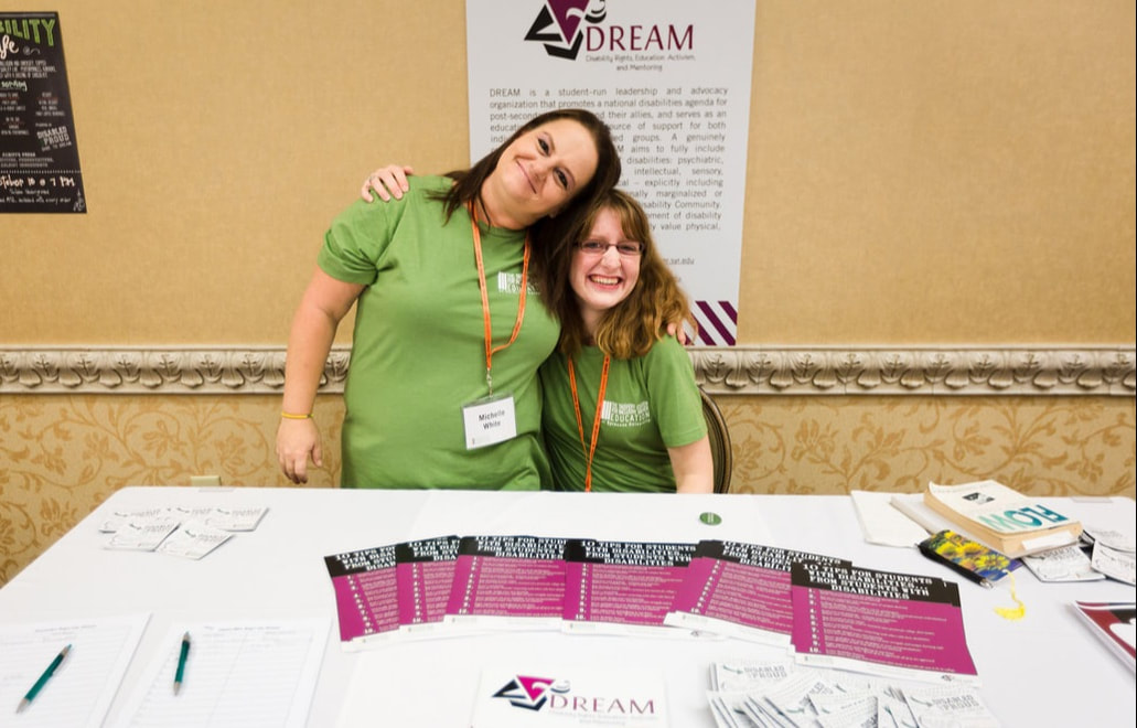Two young white women, one standing and one sitting, each wear green shirts and have a conference name badge hanging around their necks. They are smiling and each has an arm around the other. One the wall behind them is a DREAM poster and on the table in front of them are various publications and pens with the DREAM logo on them. 