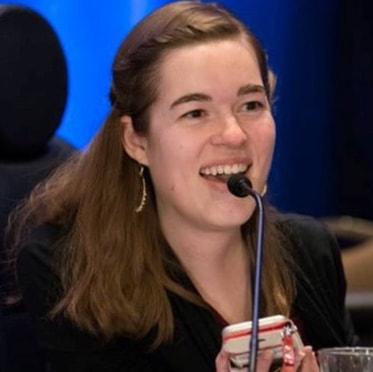 Headshot of Kings Floyd, a white woman with long dark hair. Kings, wearing a dark suit jacket and large hoop earrings, is sitting at a table with papers, notebook and water bottle in front of her. She is laughing.