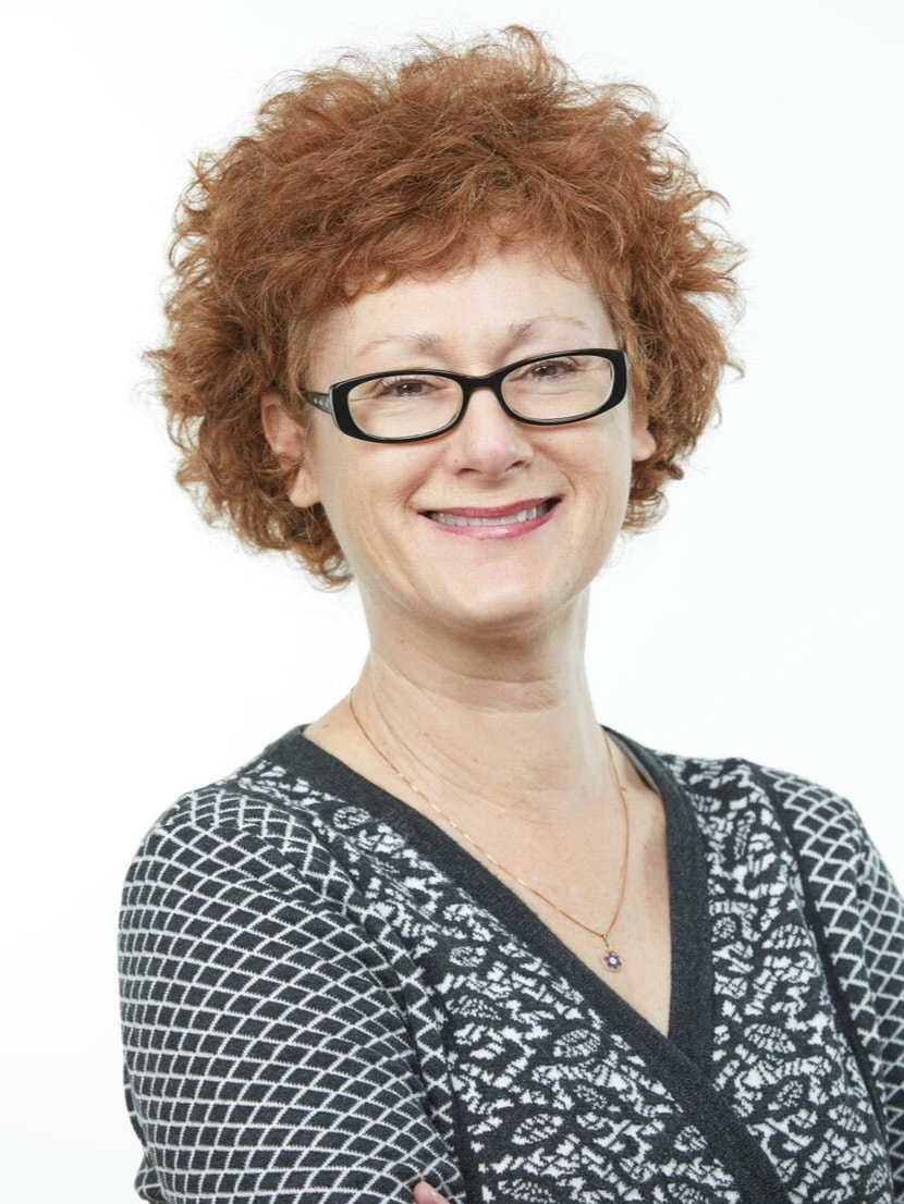 Headshot of Marci Shaffer, a white woman with short, wavy reddish brown hair and black glasses. She is wearing a black-and-white geometric print sweater and a smile.