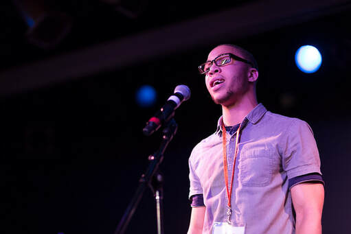 Picture of Black man iwht purple shirt standing at a microphone talking, with a dark background and bright light that implies he is on a stage
