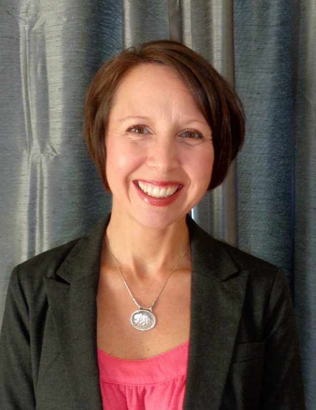 A photo of Tracie DeFreitas, a white woman with short brown hair and brown eyes. She is wearing a pink shirt and black jacket with a large silver oval pendant and smiling.